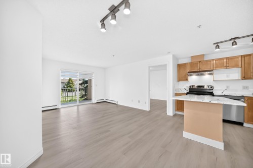 Open-concept living space featuring wood-finish flooring, a kitchen with wood cabinetry and stainless steel appliances, a breakfast bar with a light-toned countertop, and a sliding glass door leading to a balcony - #214 245 Edwards Dr Sw, Edmonton, AB - Indoor Photo Showing Kitchen