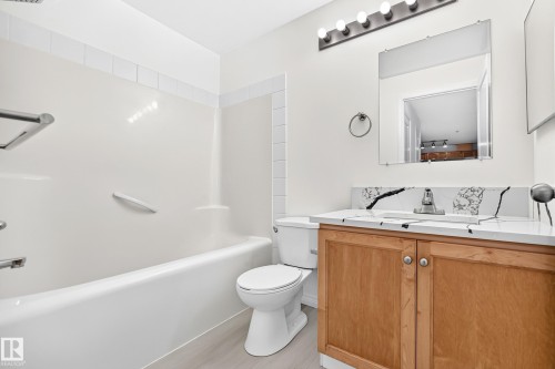 Bathroom featuring a white bathtub with an integrated shower, a toilet, and a wood-finish vanity with a white countertop and dark veining - #214 245 Edwards Dr Sw, Edmonton, AB - Indoor Photo Showing Bathroom
