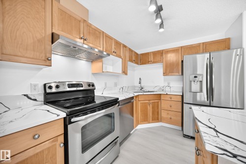 Kitchen featuring light wood cabinetry, stainless steel appliances, white countertops with prominent dark veining, and track lighting - #214 245 Edwards Dr Sw, Edmonton, AB - Indoor Photo Showing Kitchen