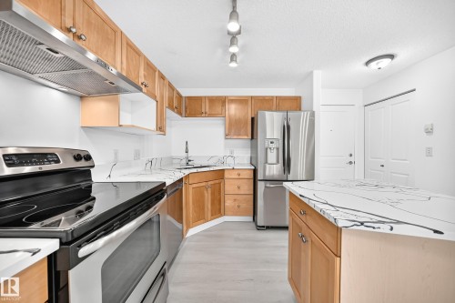 Kitchen featuring light wood cabinetry with brushed nickel hardware, white countertops with dark veining, stainless steel appliances, and light wood-finish flooring - #214 245 Edwards Dr Sw, Edmonton, AB - Indoor Photo Showing Kitchen