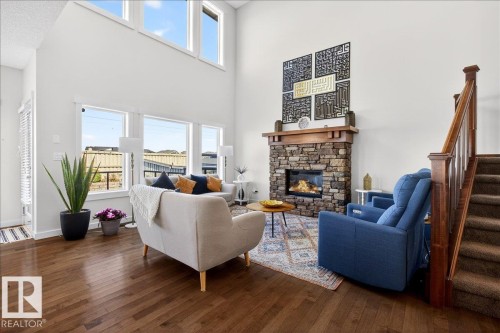 Soaring two-story great room with expansive windows flooding the space with natural light, hardwood flooring throughout, and a striking stone fireplace with built-in hearth - 3936 Claxton Loop Sw, Edmonton, AB - Indoor Photo Showing Living Room With Fireplace