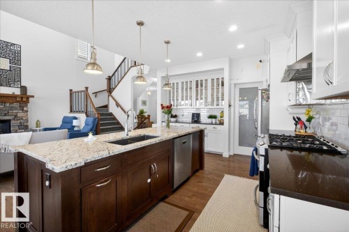 Chef's kitchen featuring a substantial dark wood island with light stone countertops, professional-grade gas range with stainless steel hood, white cabinetry with glass-front uppers, recessed lighting, and brass pendant fixtures - 3936 Claxton Loop Sw, Edmonton, AB - Indoor Photo Showing Kitchen With Double Sink With Upgraded Kitchen