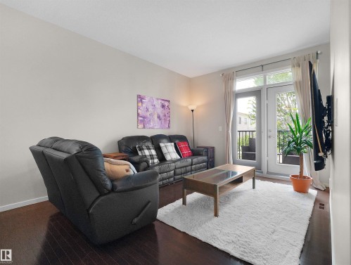 Dark wood-finish flooring, light neutral wall tones, and double French doors opening to an exterior balcony with black iron railings - #89 603 Watt Bv Sw, Edmonton, AB - Indoor Photo Showing Living Room