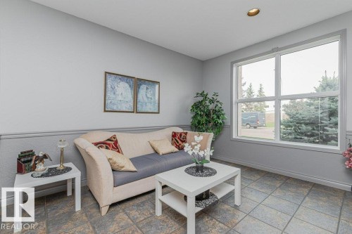 Bright corner sunroom with large multi-pane windows framing natural light, ceramic tile flooring, neutral upholstered seating area, white painted coffee table, and decorative wall art - #208 10421 42 Av Nw, Edmonton, AB - Indoor Photo Showing Living Room