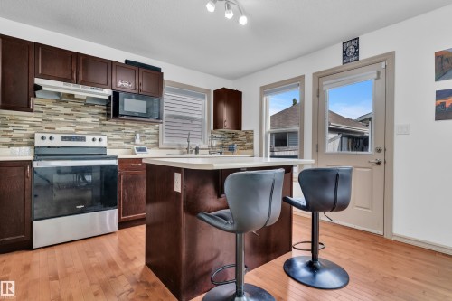 Open-concept kitchen featuring dark wood cabinetry, stainless steel appliances, a mosaic tile backsplash, a center island with countertop seating, and light wood-finish flooring - 7223 19A Avenue Sw, Edmonton, AB - Indoor Photo Showing Kitchen With Stainless Steel Kitchen