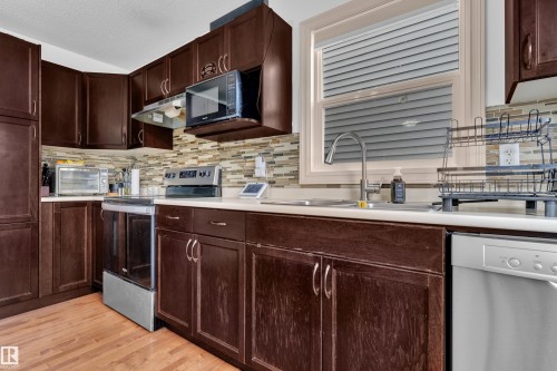 Kitchen featuring dark wood cabinetry, white countertops, a mosaic tile backsplash, stainless steel appliances, and wood-finish flooring - 7223 19A Avenue Sw, Edmonton, AB - Indoor Photo Showing Kitchen With Double Sink