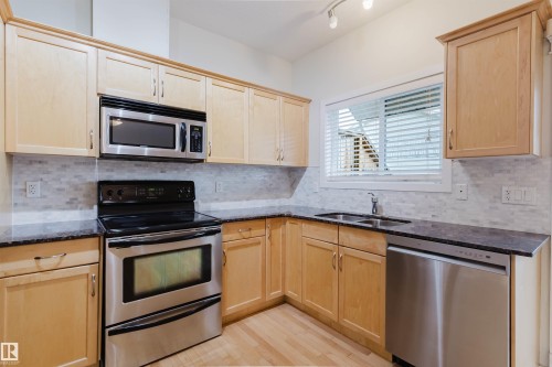Hardwood flooring, stainless steel appliances, light wood-finish cabinetry, dark countertops, and a full-height tile backsplash - #2 13215 153 Av Nw, Edmonton, AB - Indoor Photo Showing Kitchen With Double Sink