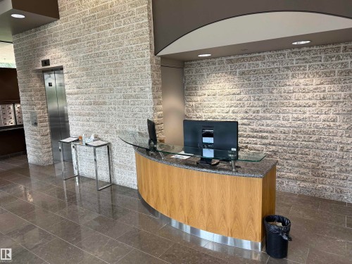 Curved wood-finish reception desk with a granite countertop, textured stone-finish accent wall, polished tile flooring, and integrated recessed lighting - #1201 10035 Saskatchewan Dr Nw, Edmonton, AB - Indoor