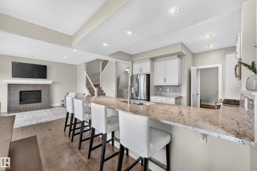 Open-concept living area featuring a kitchen island with seating, a built-in electric fireplace with tile surround, wood-finish flooring, white cabinetry, and stainless steel appliances - 1323 158 Street Sw, Edmonton, AB - Indoor Photo Showing Kitchen With Stainless Steel Kitchen With Upgraded Kitchen