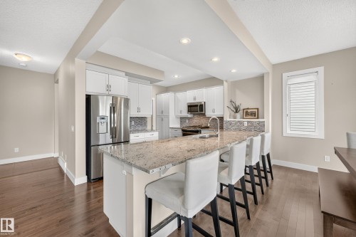 Kitchen featuring an expansive island with a light-toned countertop, white cabinetry with silver-toned hardware, stainless steel appliances, and wood-finish flooring - 1323 158 Street Sw, Edmonton, AB - Indoor Photo Showing Kitchen With Stainless Steel Kitchen With Upgraded Kitchen