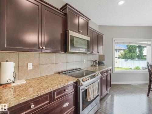 Kitchen featuring dark wood cabinetry, light-toned tiled backsplash, speckled stone countertops, and stainless steel appliances - 1478 37B Avenue Nw, Edmonton, AB - Indoor Photo Showing Kitchen With Upgraded Kitchen