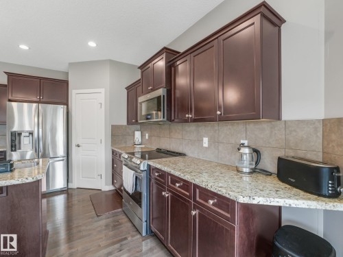 Kitchen featuring rich wood cabinetry, granite-style countertops, stainless steel appliances, tile backsplash, and wood-finish flooring - 1478 37B Avenue Nw, Edmonton, AB - Indoor Photo Showing Kitchen With Upgraded Kitchen