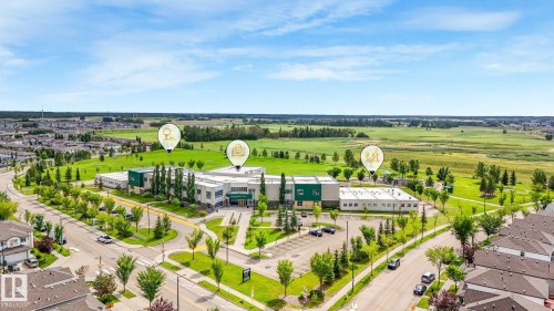 Aerial perspective of a developed neighborhood bordering open green spaces, featuring a large institutional building with a parking lot, paved roadways, and residential rooftops - 1478 37B Avenue Nw, Edmonton, AB - Outdoor With View