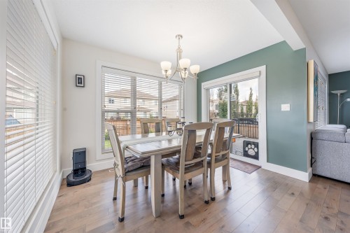 Wood-finish flooring throughout, multiple large windows with white blinds, contemporary chandelier, sliding glass door, and a painted accent wall - 76 Dunlop Wynd, Leduc, AB - Indoor Photo Showing Dining Room