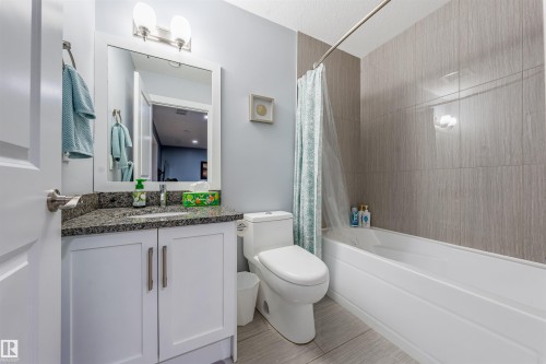 Modern bathroom featuring a white vanity with a granite countertop, a rectangular mirror, a shower-over-tub combination with textured wall tiling, and a white toilet - 76 Dunlop Wynd, Leduc, AB - Indoor Photo Showing Bathroom