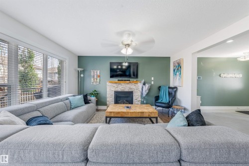 Expansive living space featuring a stone-clad fireplace with wood mantle, large windows with blinds, and a ceiling fan - 76 Dunlop Wynd, Leduc, AB - Indoor Photo Showing Living Room With Fireplace