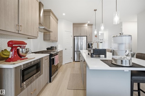 Modern kitchen featuring wood-finish cabinetry, stainless steel appliances, a white herringbone backsplash, and a large island with pendant lighting - 3877 Chrustawka Place Sw, Edmonton, AB - Indoor Photo Showing Kitchen With Upgraded Kitchen