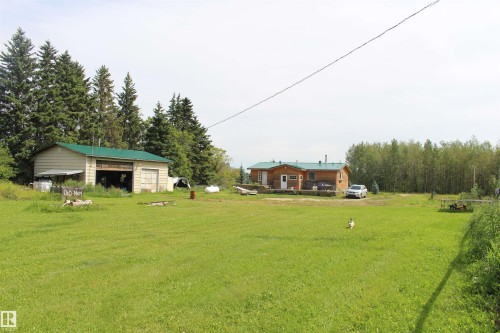 Expansive green lawn, detached garage with dual bay doors, wood-sided residence with green roof, mature evergreen trees, and additional surrounding foliage - 49480 Rge Rd 41, Rural Leduc County, AB - Outdoor