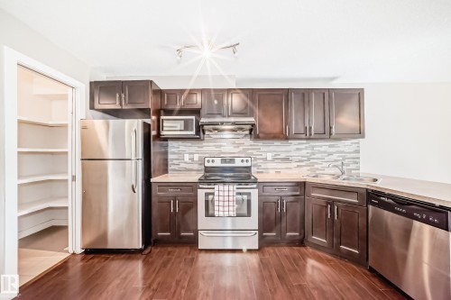 Kitchen featuring stainless steel appliances, dark wood-finish cabinetry, light-toned countertops, a full mosaic tile backsplash, and wood-finish flooring - 12118 125 Street Nw, Edmonton, AB - Indoor Photo Showing Kitchen