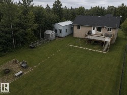 Expansive backyard featuring a large elevated deck with stairs, a stone patio area with a fire pit, a detached outbuilding, and a separate shed with raised garden beds, all surrounded by mature trees - 