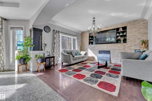 Hardwood flooring, natural light through large windows, a stacked stone accent wall with integrated shelving, a built-in fireplace, and an elegant chandelier - 10915 175 Avenue Nw, Edmonton, AB - Indoor Photo Showing Living Room With Fireplace