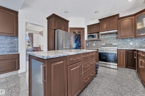 Kitchen featuring wood-finish cabinetry, stainless steel appliances, a multi-tone tile backsplash, a center island with a light-toned countertop, and speckled flooring - 10915 175 Avenue Nw, Edmonton, AB - Indoor Photo Showing Kitchen With Upgraded Kitchen