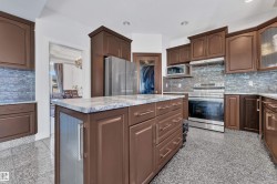 Kitchen featuring wood-finish cabinetry, stainless steel appliances, a multi-tone tile backsplash, a center island with a light-toned countertop, and speckled flooring - 