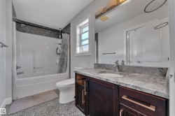 Bathroom featuring a single vanity with an integrated sink, stone countertop, dark wood-finish cabinetry with bar pull hardware, and a tub/shower combination with a textured tile surround - 