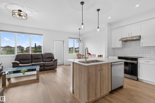 Open-concept living space featuring wood-finish flooring, white cabinetry, a kitchen island with seating, stainless steel appliances, and large windows - 1676 27 Street Nw, Edmonton, AB 
