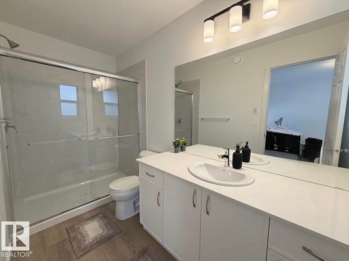 Glass-enclosed shower with chrome fixtures, wood-finish flooring, white vanity with integrated sink, expansive mirror, and modern three-light fixture - 1676 27 Street Nw, Edmonton, AB 