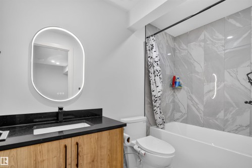 Bathroom featuring a white soaking tub, large format tile surround, a wood-finish vanity with a dark countertop, and an illuminated oval mirror - 1676 27 Street Nw, Edmonton, AB 