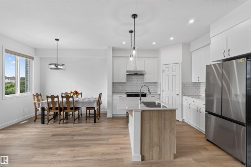 Open-concept kitchen and dining area featuring wood-finish flooring, white cabinetry, a kitchen island with sink, stainless steel appliances, and subway tile backsplash - 1676 27 Street Nw, Edmonton, AB 