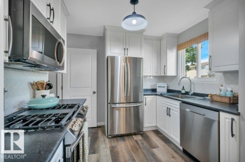 Kitchen featuring moden white cabinetry, stainless steel appliances, vinyl plank flooring, dark countertops, and  modern tile backsplash - #138 5231 51 St, Bon Accord, AB - Indoor Photo Showing Kitchen