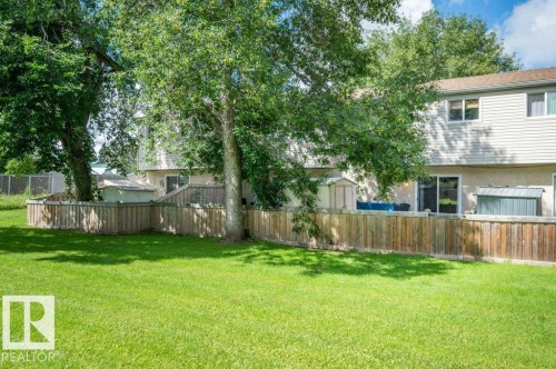 Expansive green space with a wooden privacy fence, mature trees, and a sliding glass door entry to the building - #138 5231 51 St, Bon Accord, AB - Outdoor With Backyard