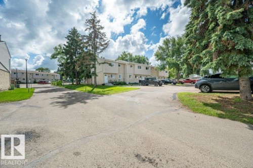 Paved parking area surrounded by mature trees and grass, with multi-unit structures in the background - #138 5231 51 St, Bon Accord, AB - Outdoor