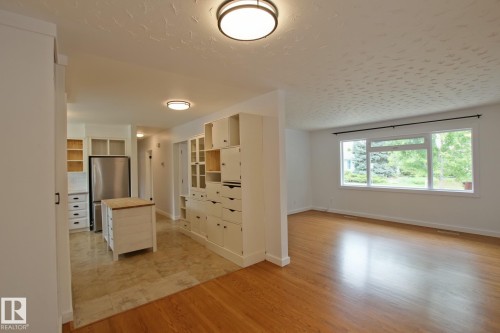 Spacious interior with wood-finish flooring, a large window, a built-in storage unit, a kitchen island, and a stainless steel refrigerator - 5712 113B Street Nw, Edmonton, AB - Indoor Photo Showing Other Room