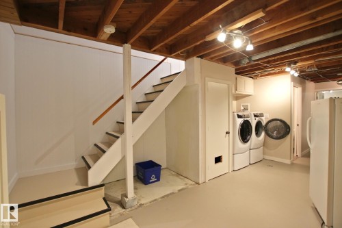 Exposed wood-beam ceiling with track lighting, interior staircase with wood handrails, painted concrete flooring, white paneled walls, and built-in laundry area - 5712 113B Street Nw, Edmonton, AB - Indoor Photo Showing Basement