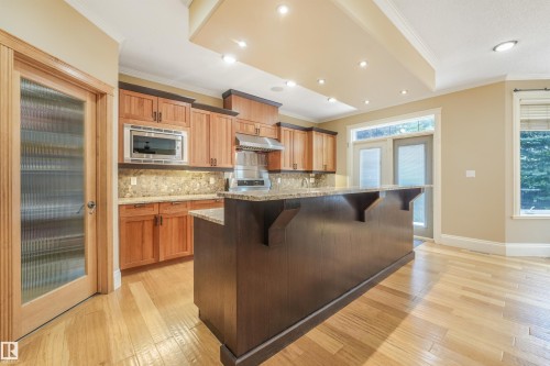 Kitchen featuring wood cabinetry, a large center island with a granite countertop, a stone-tiled backsplash, stainless steel appliances, and wood-finish flooring - 27 Kingdom Place, Leduc, AB - Indoor Photo Showing Kitchen