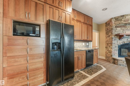 Built-in microwave, integrated black appliances, wood-finish cabinetry, stone fireplace, and patterned floor tile - 27 Kingdom Place, Leduc, AB - Indoor With Fireplace