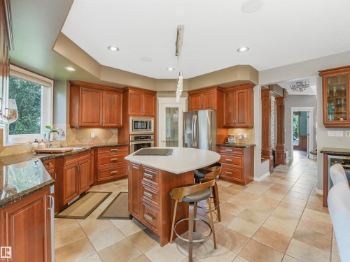 Spacious kitchen featuring custom wood cabinetry, stone countertops, a large center island with seating, stainless steel appliances, and tile flooring - 967 Lamb Crescent Nw, Edmonton, AB - Indoor Photo Showing Kitchen