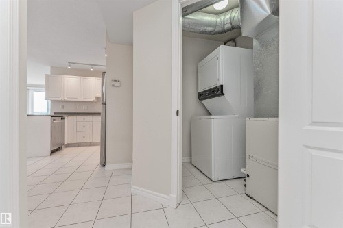 White tile flooring throughout, with a stacked washer and dryer unit, and a kitchen featuring white cabinetry, stainless steel appliances, and dark countertops - #1101 10909 103 Av Nw, Edmonton, AB - Indoor Photo Showing Laundry Room