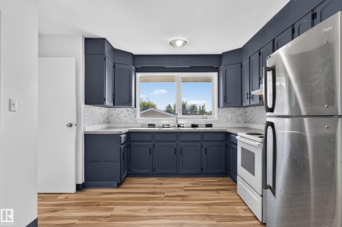 U-shaped kitchen featuring wood-finish flooring, dark-toned cabinetry, light countertops with a patterned backsplash, and a stainless steel refrigerator - 13903 87 Street Nw, Edmonton, AB - Indoor Photo Showing Kitchen With Double Sink