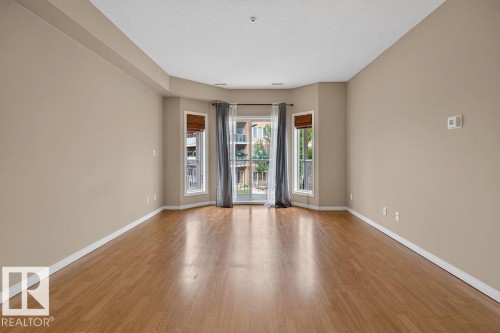 Wood-finish flooring throughout, light neutral wall paint, white ceiling, and a bay window configuration with a glass-paneled door - #205 4304 139 Av Nw, Edmonton, AB - Indoor Photo Showing Other Room