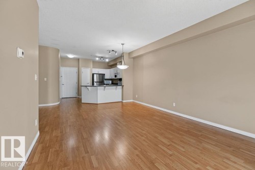 Wood-finish flooring throughout, curved wall detail, kitchen with white cabinetry, stainless steel appliances, and a breakfast bar - #205 4304 139 Av Nw, Edmonton, AB - Indoor Photo Showing Kitchen