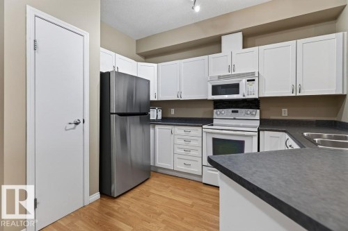 Kitchen featuring wood-finish flooring, white cabinetry, dark countertops, a double basin sink, and stainless-steel appliances - #205 4304 139 Av Nw, Edmonton, AB - Indoor Photo Showing Kitchen With Double Sink