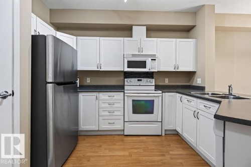 Kitchen featuring white shaker-style cabinetry, dark countertops, wood-finish flooring, and stainless steel appliances including a refrigerator, range, microwave, and double sink - #205 4304 139 Av Nw, Edmonton, AB - Indoor Photo Showing Kitchen With Double Sink