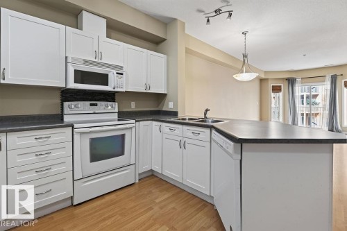 White cabinetry with stainless steel hardware, built-in microwave, wood-finish flooring, and a double basin sink - #205 4304 139 Av Nw, Edmonton, AB - Indoor Photo Showing Kitchen With Double Sink