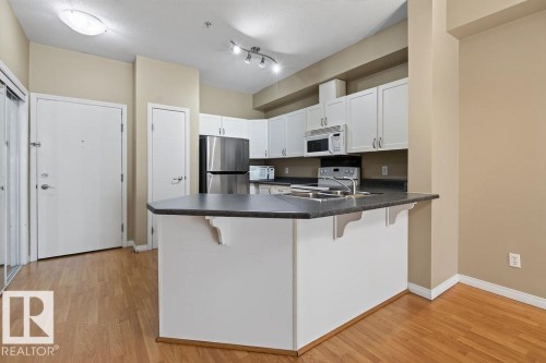 Kitchen featuring white cabinetry, stainless steel appliances, dark countertops, and wood-finish flooring - #205 4304 139 Av Nw, Edmonton, AB - Indoor Photo Showing Kitchen
