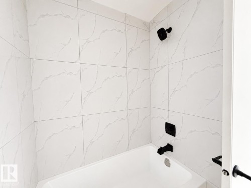 Bathtub featuring a white finish, large format wall tiling with subtle veining, matte black showerhead, and matte black bath fixtures - 13118 186 Avenue Nw, Edmonton, AB - Indoor Photo Showing Bathroom