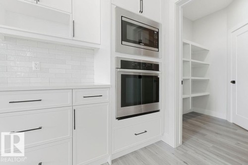 White shaker-style cabinetry with matte black hardware, stainless steel built-in microwave and oven, white subway tile backsplash, light wood-finish flooring, and a walk-in pantry with built-in shelving - 9331 181 Avenue Nw, Edmonton, AB - Indoor